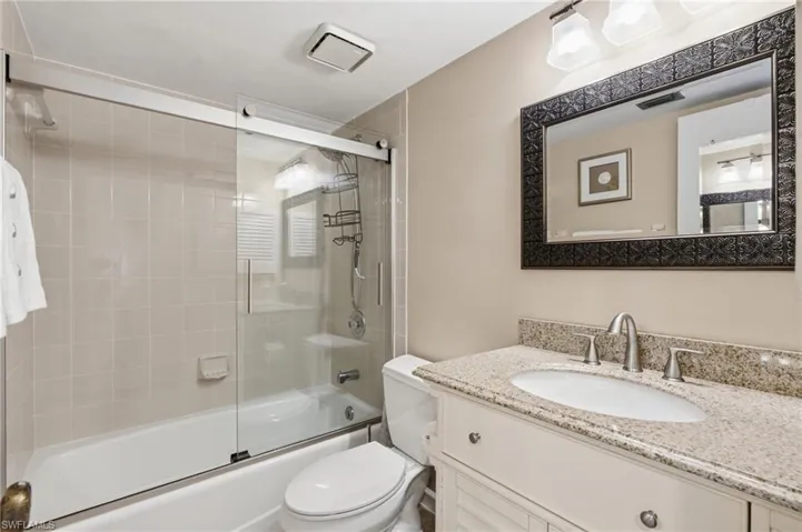Bathroom featuring a combination tub and shower with glass sliding doors, neutral-toned wall tiles, and a white vanity with a light-colored countertop - Virtually Edited Image