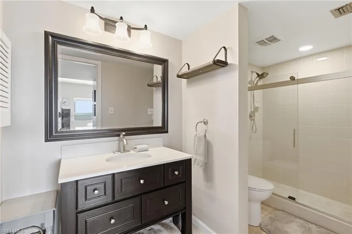 Bathroom featuring a dark wood vanity with a white countertop, framed mirror, three-light fixture, and a glass-enclosed shower with tiled walls - Virtually Edited Image