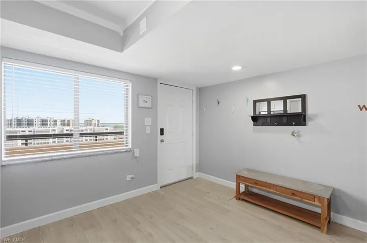 Bright entry area featuring a large window with horizontal blinds, light wood-finish flooring, a white paneled door, recessed lighting, and white baseboards - Virtually Edited Image