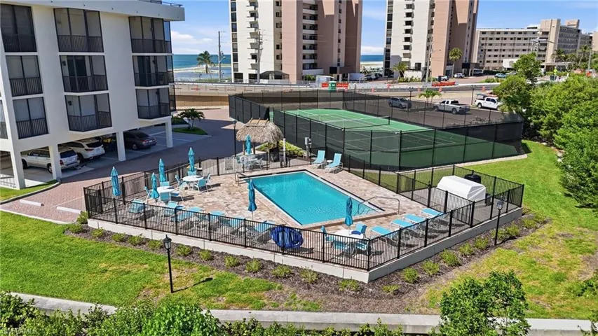 Outdoor swimming pool area with paver decking, black metal perimeter fencing, and a nearby tennis court - Virtually Edited Image