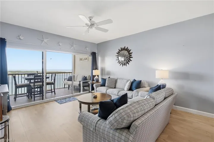 Spacious living area featuring light wood-finish flooring, a ceiling fan, and direct access to a private balcony through a large sliding glass door - Virtually Edited Image