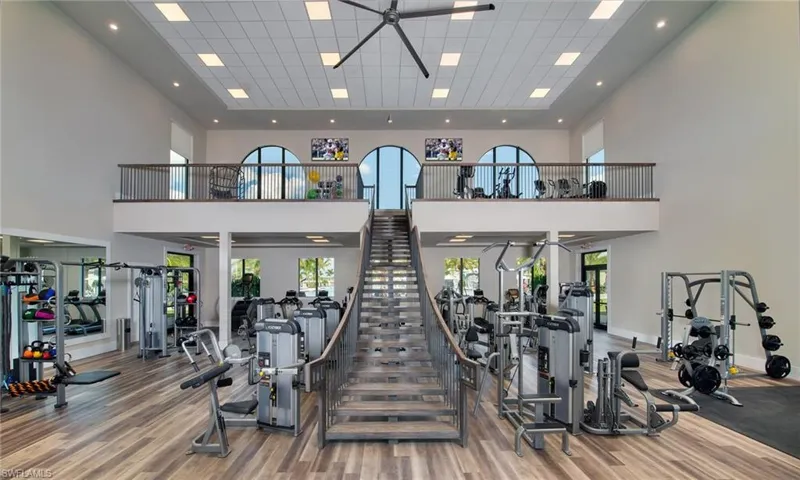 High-ceiling fitness area featuring wood-finish flooring, a grand staircase with metal railings, and a second-story fitness loft with arched windows