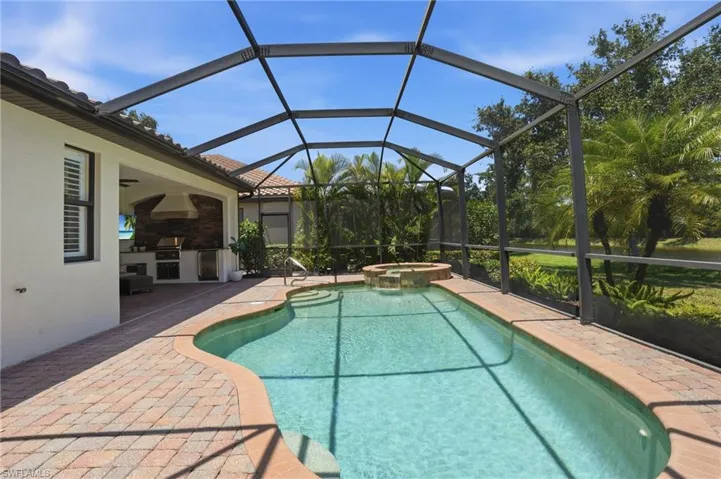 Screened outdoor living area featuring a custom-shaped pool with integrated spa, paver patio, full outdoor kitchen with built-in grill, and mature landscaping
