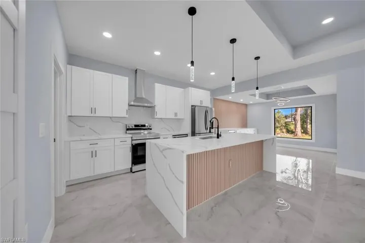 Sleek kitchen featuring white shaker cabinetry, stainless steel appliances, a large center island with a fluted wood-finish panel, quartz-finish countertops and backsplash, and contemporary pendant lighting