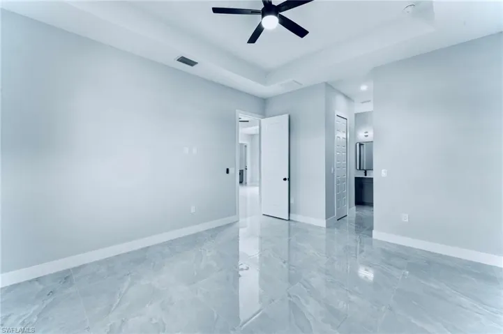 Unfurnished bedroom featuring a tray ceiling, ensuite bath, light marble finish flooring, ceiling fan, and a closet
