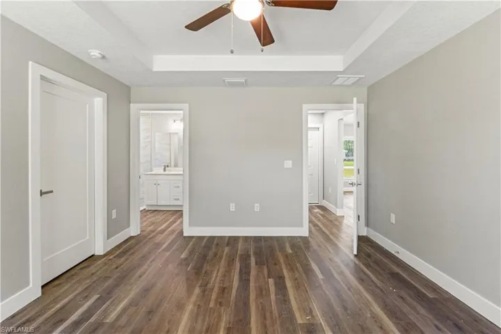 Unfurnished bedroom with a tray ceiling, ensuite bath, dark wood-style floors, ceiling fan, and a textured ceiling
