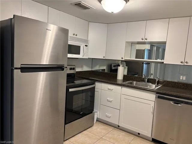 Kitchen with sink, light tile patterned floors, dark stone counters, white cabinets, and appliances with stainless steel finishes
