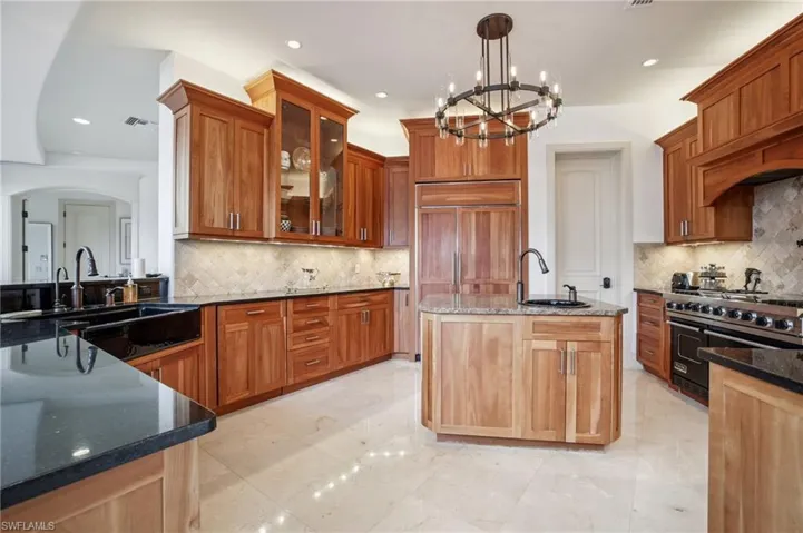 Kitchen with dark stone countertops, a chandelier, tasteful backsplash, a kitchen island with sink, and pendant lighting