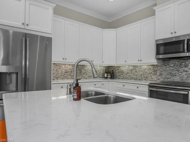 Kitchen with decorative backsplash, appliances with stainless steel finishes, light quartz countertops, ornamental molding, and white cabinets.