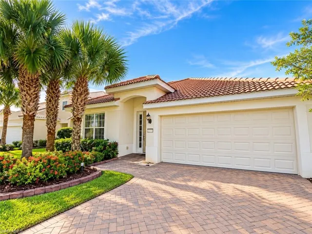 Stucco exterior featuring a tiled roof, multi-car garage, paver driveway, and landscaped garden beds with mature palm trees