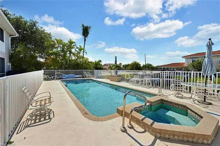 Outdoor swimming pool and spa with a concrete deck, white perimeter fencing, and established tropical landscaping