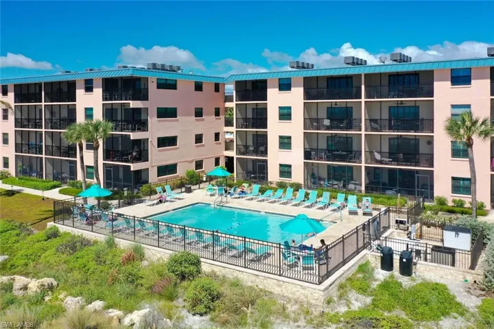 Multi-story building complex featuring private screened balconies, ground-level swimming pool, metal perimeter fencing, and established palm landscaping