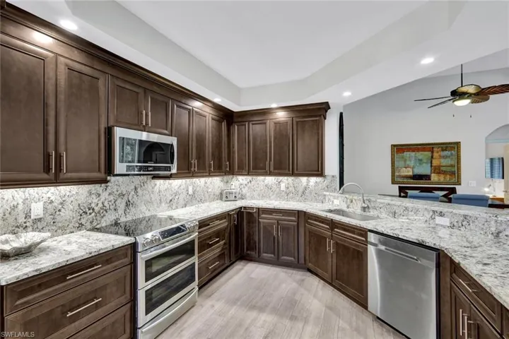 Kitchen featuring dark wood cabinetry, light-toned granite countertops and backsplash, stainless steel appliances, under-cabinet lighting, and wood-finish flooring