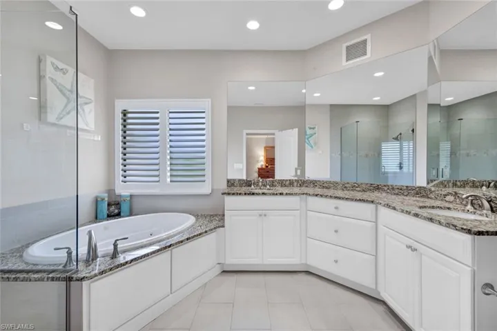 Bathroom featuring a built-in jetted tub with granite surround, white plantation shutters, dual vanities with granite countertops, and a glass-enclosed shower