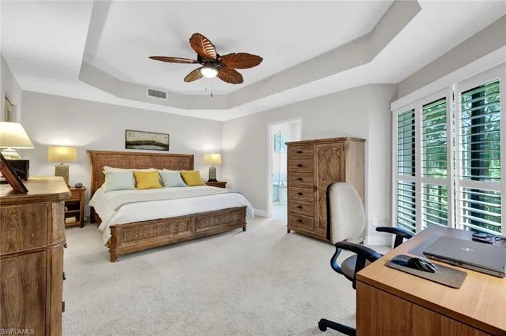 Spacious primary bedroom featuring a tray ceiling with a ceiling fan,  large windows with plantation shutters, and an open doorway leading to an adjacent bathroom