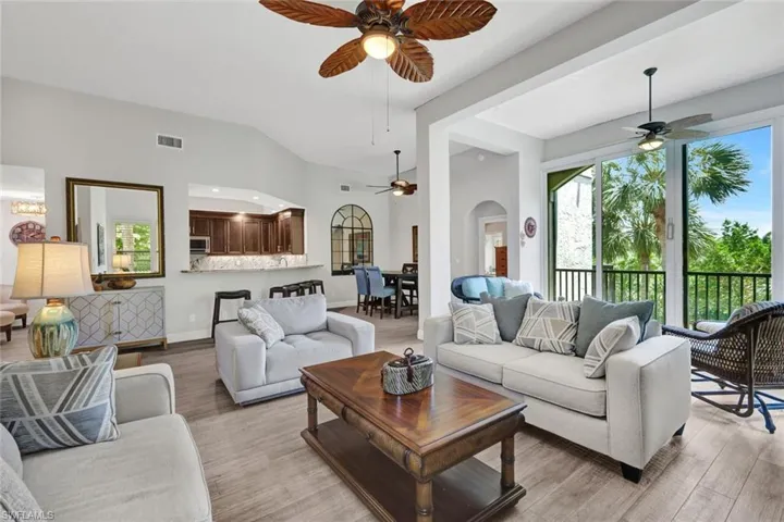 Vaulted ceilings and wood-finish flooring define this open living space, featuring a kitchen pass-through with a breakfast bar, a dining area beyond, and sliding glass doors opening to a balcony with black railings