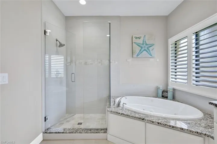 Spacious bathroom featuring a frameless glass shower enclosure, a built-in soaking tub, plantation shutters, a tiled shower with a decorative accent band, and a granite-topped tub surround