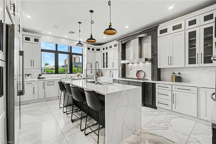 Kitchen featuring an expansive center island with waterfall countertop, integrated gas range with stainless steel hood, white and dark wood-finish cabinetry, and large picture windows