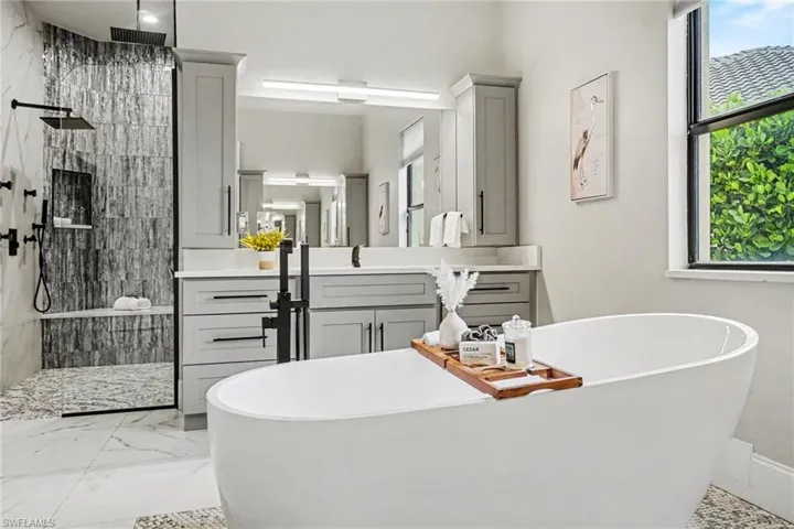 Freestanding soaking tub with a floor-mounted matte black faucet, walk-in shower with a built-in bench and rain shower head, dual vanity with quartz countertops and undermount sinks, and large-format tile flooring