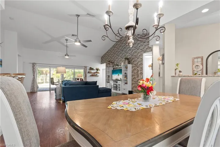 The dining area features a wooden table with a classic chandelier overhead, and offers a view of the living area with vaulted ceilings, dark wood flooring, and sliding glass doors that open to the outdoor space