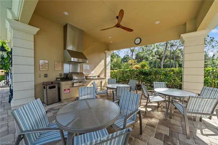 The community features a covered outdoor patio area with a built-in BBQ grill, an overhead fan, and ample seating with tables and chairs, all set on a paver stone patio