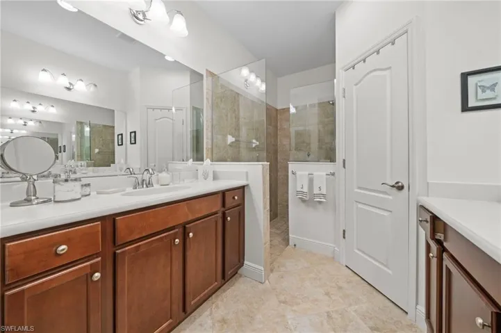 Bathroom featuring a double vanity with brown cabinetry and white countertops, a tiled walk-in shower with a glass enclosure, and light-colored tiled flooring