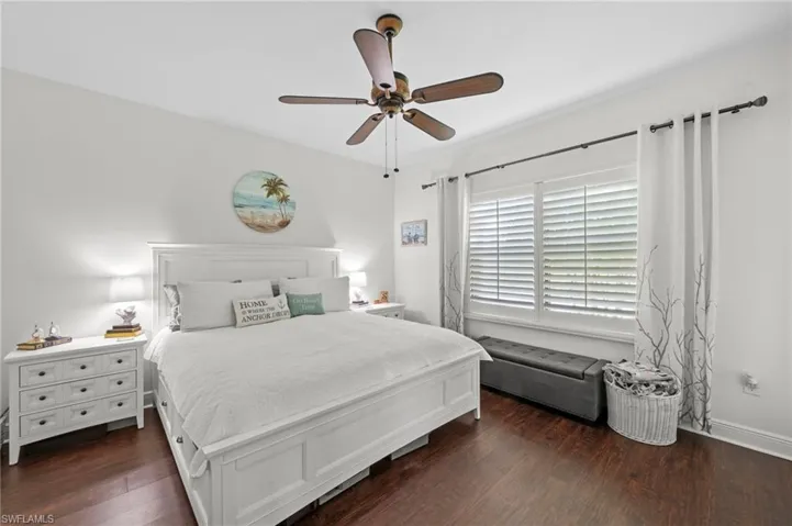The bedroom features dark wood flooring, a ceiling fan with a light fixture, and a window fitted with white shutters and light-colored curtains