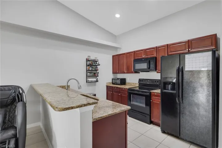 Kitchen featuring black appliances, a peninsula, reddish brown cabinets, light stone countertops, and vaulted ceiling