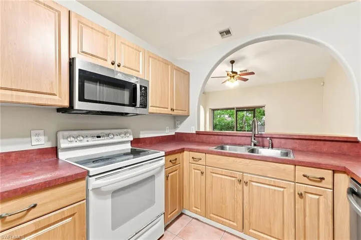 Kitchen featuring light wood cabinetry, a stainless steel over-the-range microwave, a white electric range, a double basin stainless steel sink, and red countertops