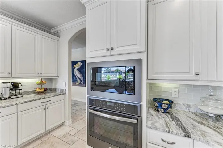 Kitchen with stainless steel oven, backsplash, light stone counters, and white cabinets
