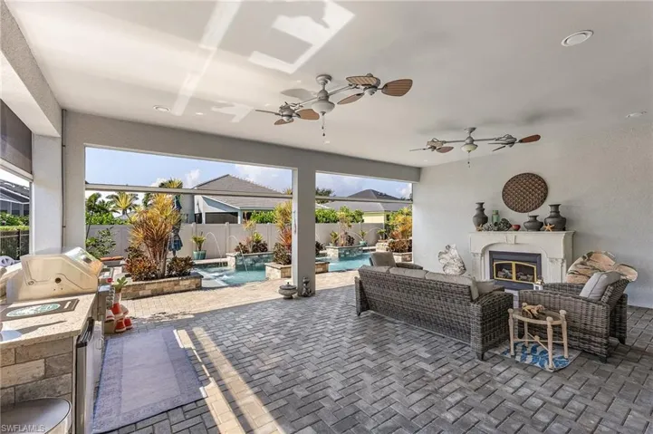 Covered outdoor living area featuring paver flooring, a built-in outdoor kitchen with stone veneer, a fireplace with a decorative mantle, and dual ceiling fans