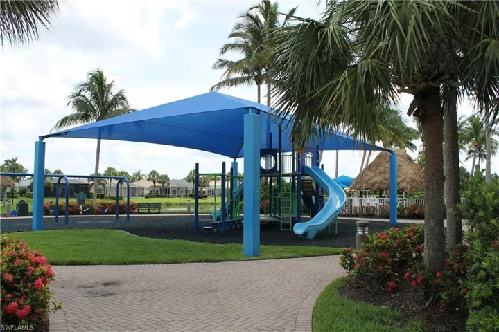 Outdoor recreation area featuring a shaded playground structure, spiral slide, paved brick pathways, and mature palm trees