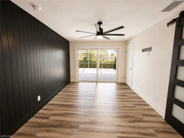 Wood-finish flooring, accent wall with vertical paneling, contemporary ceiling fan, white sliding glass doors, and a dark-toned barn door with frosted glass panels