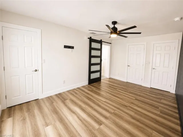 Wood-finish flooring with white baseboards, featuring a contemporary ceiling fan with integrated lighting, a modern barn door with frosted glass panels, and white paneled doors