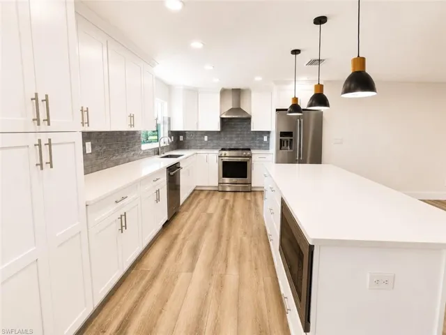 White cabinetry, light wood-finish flooring, stainless steel appliances, dark gray subway tile backsplash, and a large central island