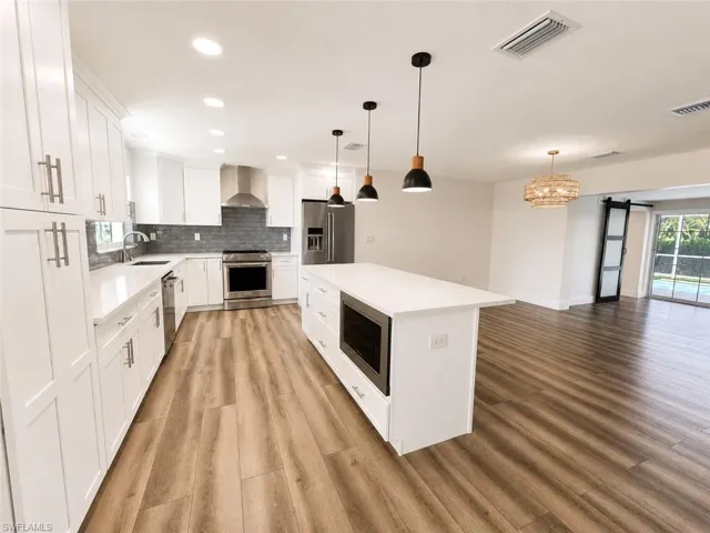 White cabinetry with brushed nickel hardware, stainless steel appliances, wood-finish flooring, a kitchen island with built-in microwave, and pendant lighting
