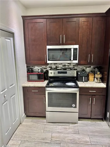 Kitchen featuring appliances with stainless steel finishes, decorative backsplash, light stone counters, dark brown cabinets, and a textured wall