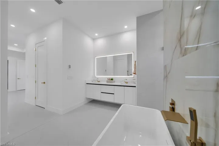 Freestanding soaking tub with brushed gold fixtures, a floating dual vanity with an illuminated mirror, large format wall tiles with subtle veining, recessed lighting, and light-toned flooring