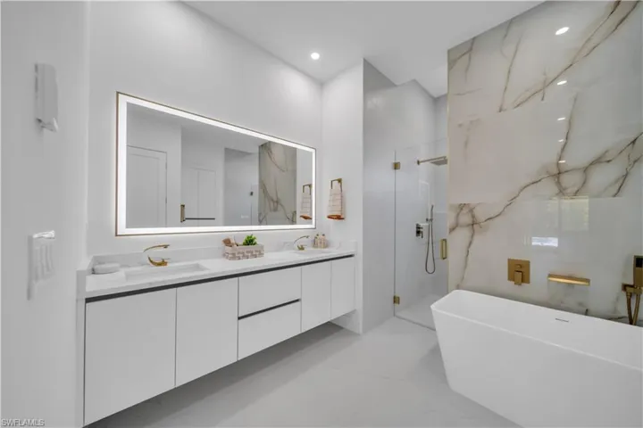 Modern bathroom featuring a dual vanity with white cabinetry, gold-finish hardware, and a lighted mirror