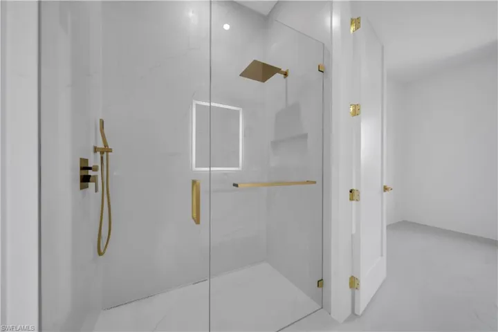 Contemporary glass-enclosed shower featuring large format white tile with subtle veining, a rainfall showerhead, handheld sprayer, and built-in shelving, all with gold-tone fixtures