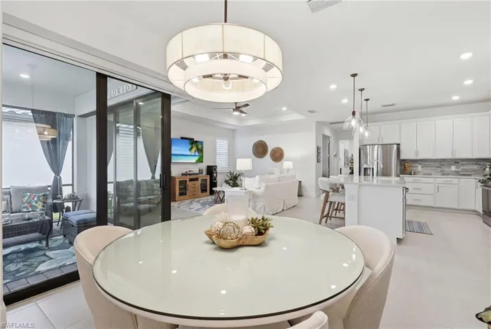Modern open-concept interior featuring light-toned flooring, a round glass-top dining table, a kitchen island with pendant lighting, white cabinetry, and stainless steel appliances