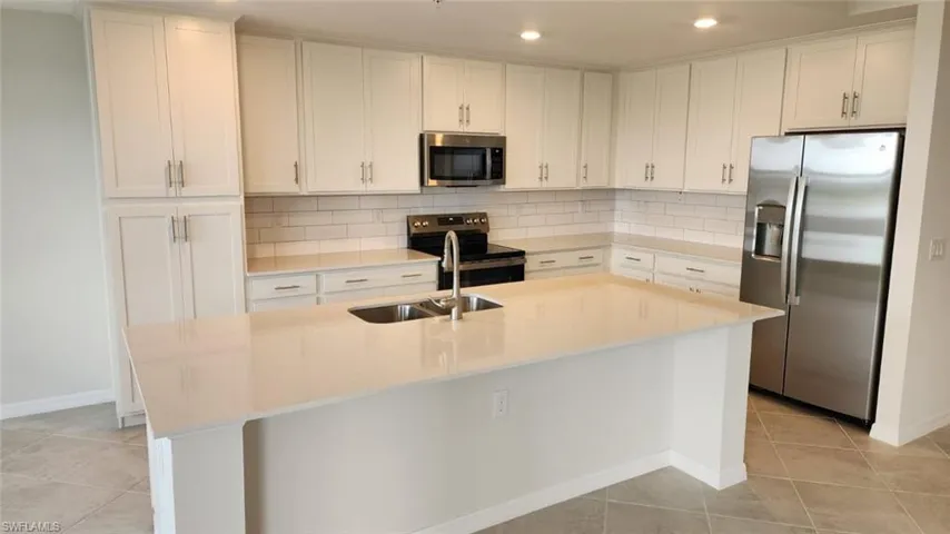 Kitchen featuring white shaker-style cabinetry, a spacious island with a double basin sink, stainless steel appliances, subway tile backsplash, and light-toned countertops