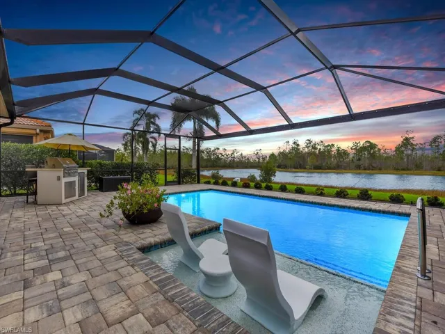Pool at dusk with glass enclosure, a patio area, a water view, and exterior kitchen