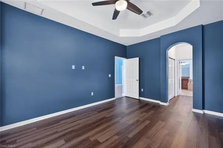 Unfurnished bedroom with arched walkways, a tray ceiling, ensuite bathroom, dark wood finished floors, and ceiling fan