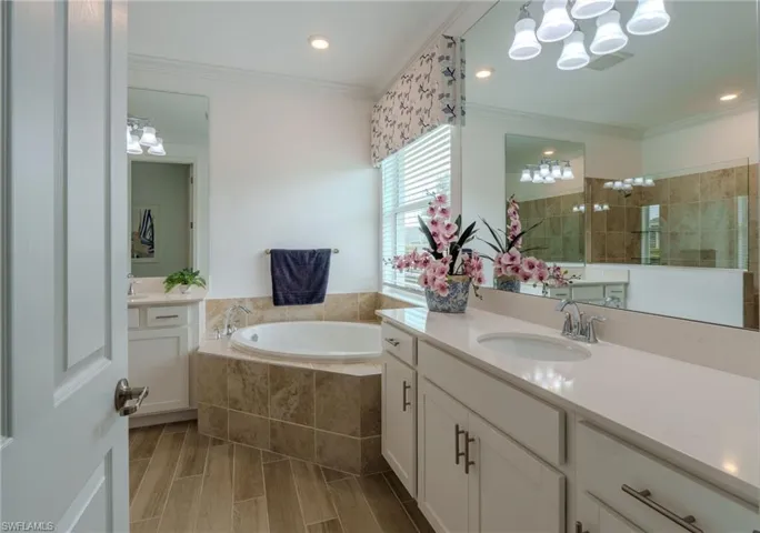 Spacious bathroom featuring a built-in soaking tub with stone tile surround, dual vanities with quartz countertops, wood-finish flooring, and a large mirror