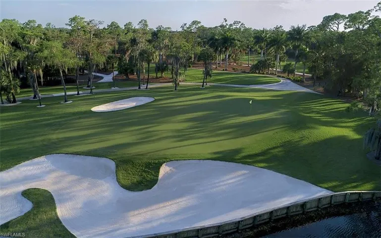 Expansive golf course green with multiple sand traps, surrounded by mature trees, and bordered by a water feature with a retaining wall
