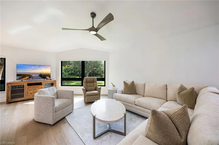 Bright, open living room featuring hardwood flooring, a ceiling fan with brushed metal finish, and expansive windows framed by black mullions overlooking mature landscaping