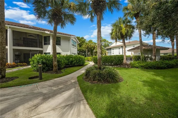 Concrete pathway with lush green lawn, bordered by mature palm trees and manicured hedges, leading to a multi-story building with a tiled roof and screened balconies