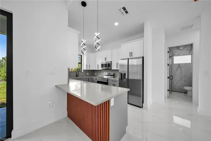 Kitchen with appliances with stainless steel finishes, a peninsula, white cabinetry, light marble finish floors, and decorative light fixtures