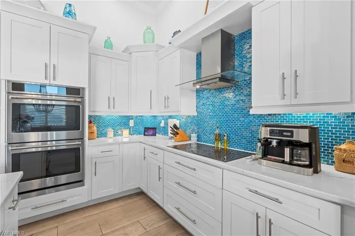 Kitchen featuring wall chimney range hood, black electric stovetop, light stone countertops, white cabinets, and stainless steel double oven